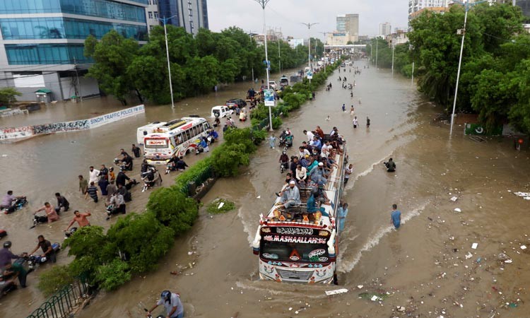 People sit atop a bus roof while others wade through the flooded road during monsoon rain in Karachi